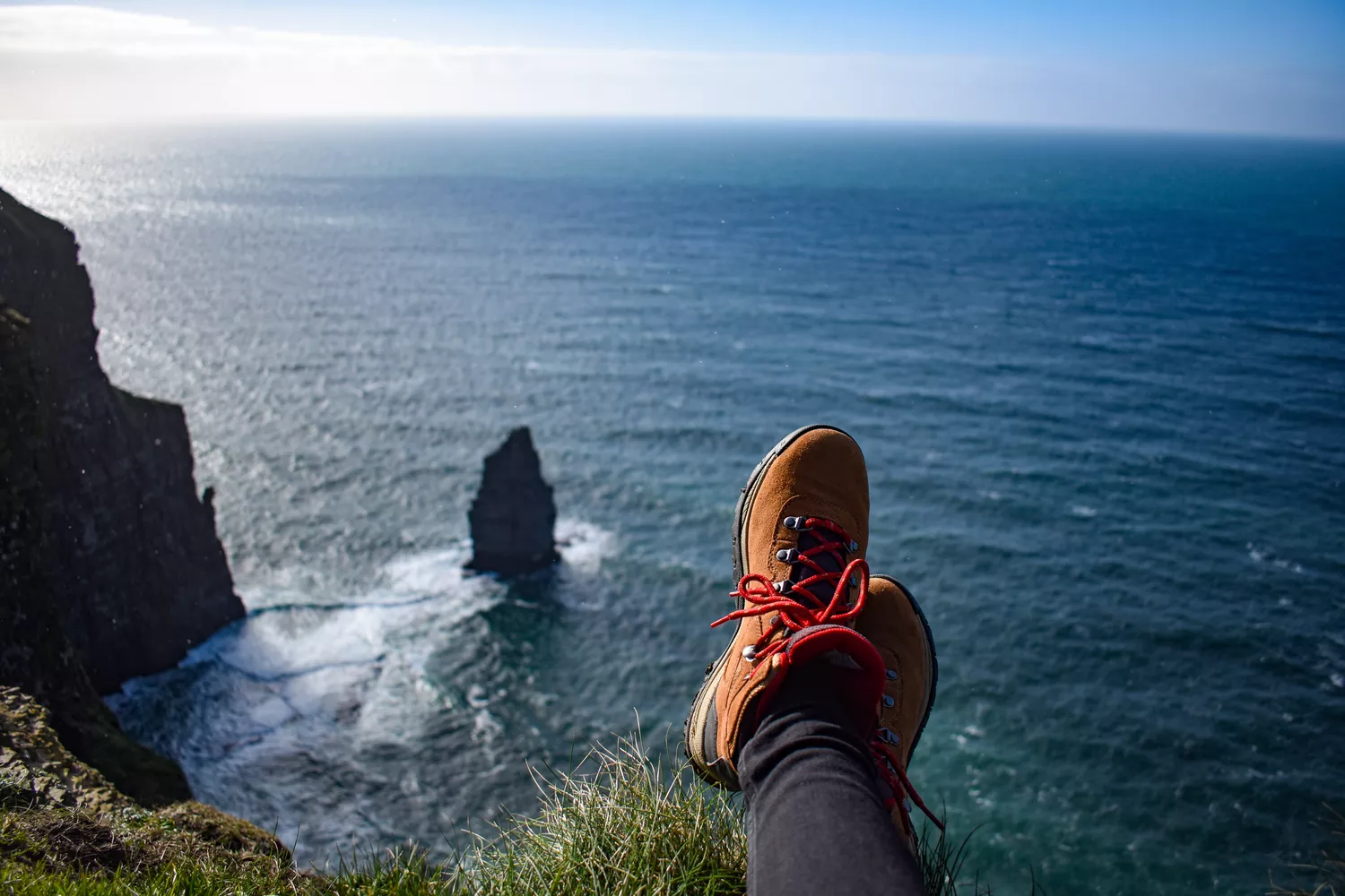 A person's feet with hiking boots on perched on a cliff's edge, gazing out at the vast, sparkling ocean beneath.